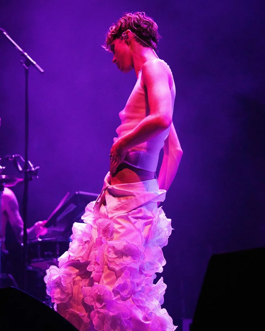 A person in a white top and ruffled floral skirt stands on stage with a microphone and keyboard in the background, illuminated by purple lighting.