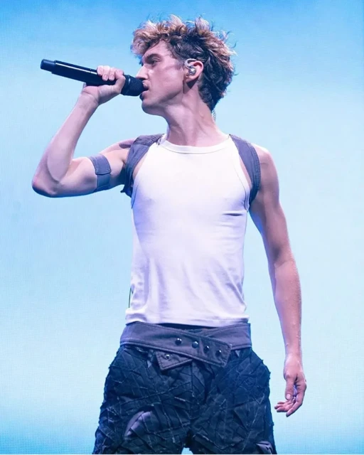 Young male singer performs on stage with a microphone, wearing a white tank top and black textured pants under bright concert lighting.