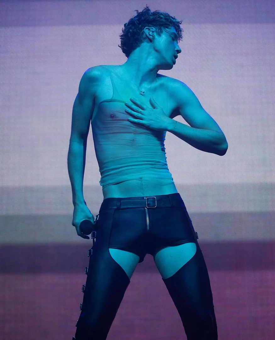 A singer performs on stage under blue lighting wearing black chaps, a sheer top and holding a microphone in hand.
