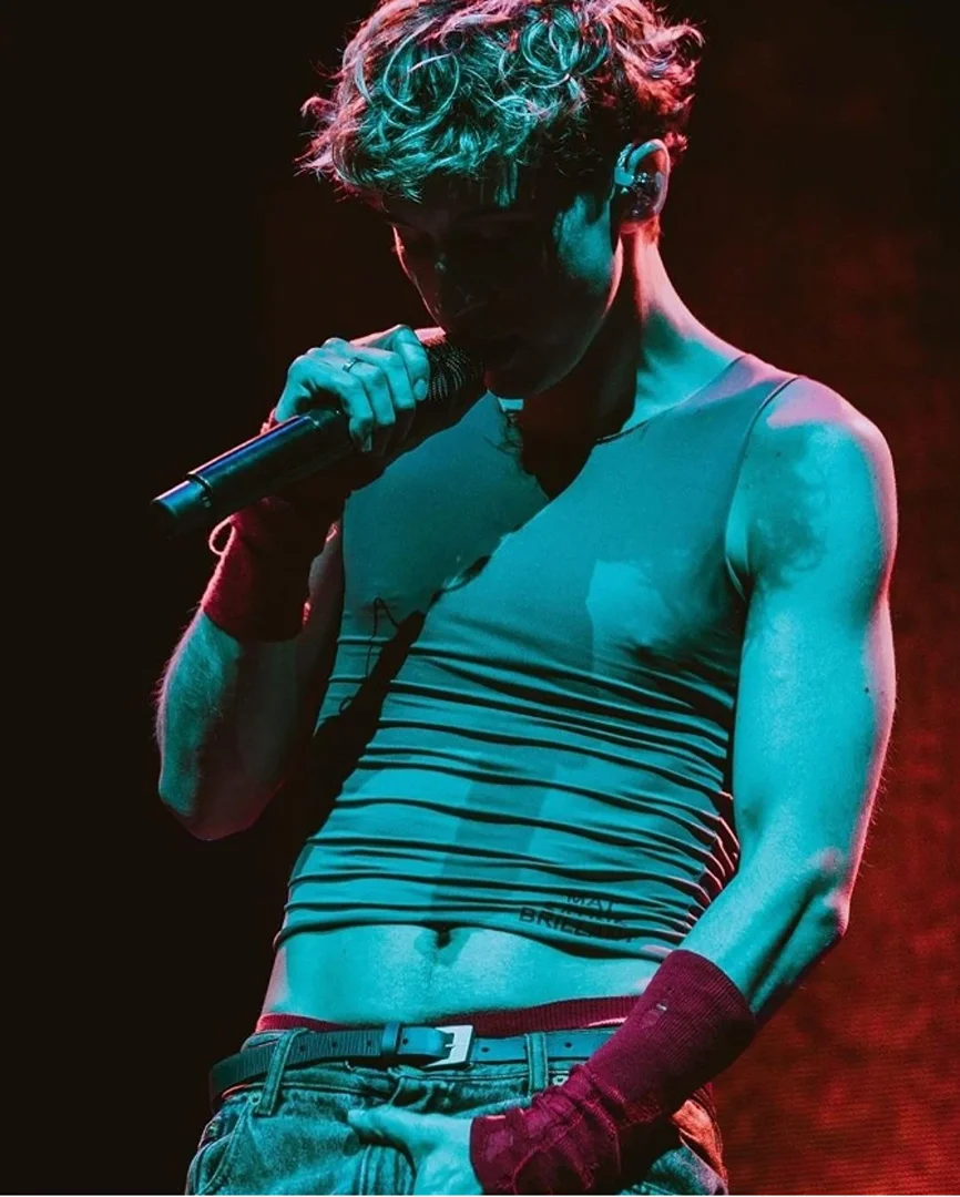 Man singing on stage under vibrant lighting, wearing a tank top and jeans, holding a microphone with his hand, performing live.