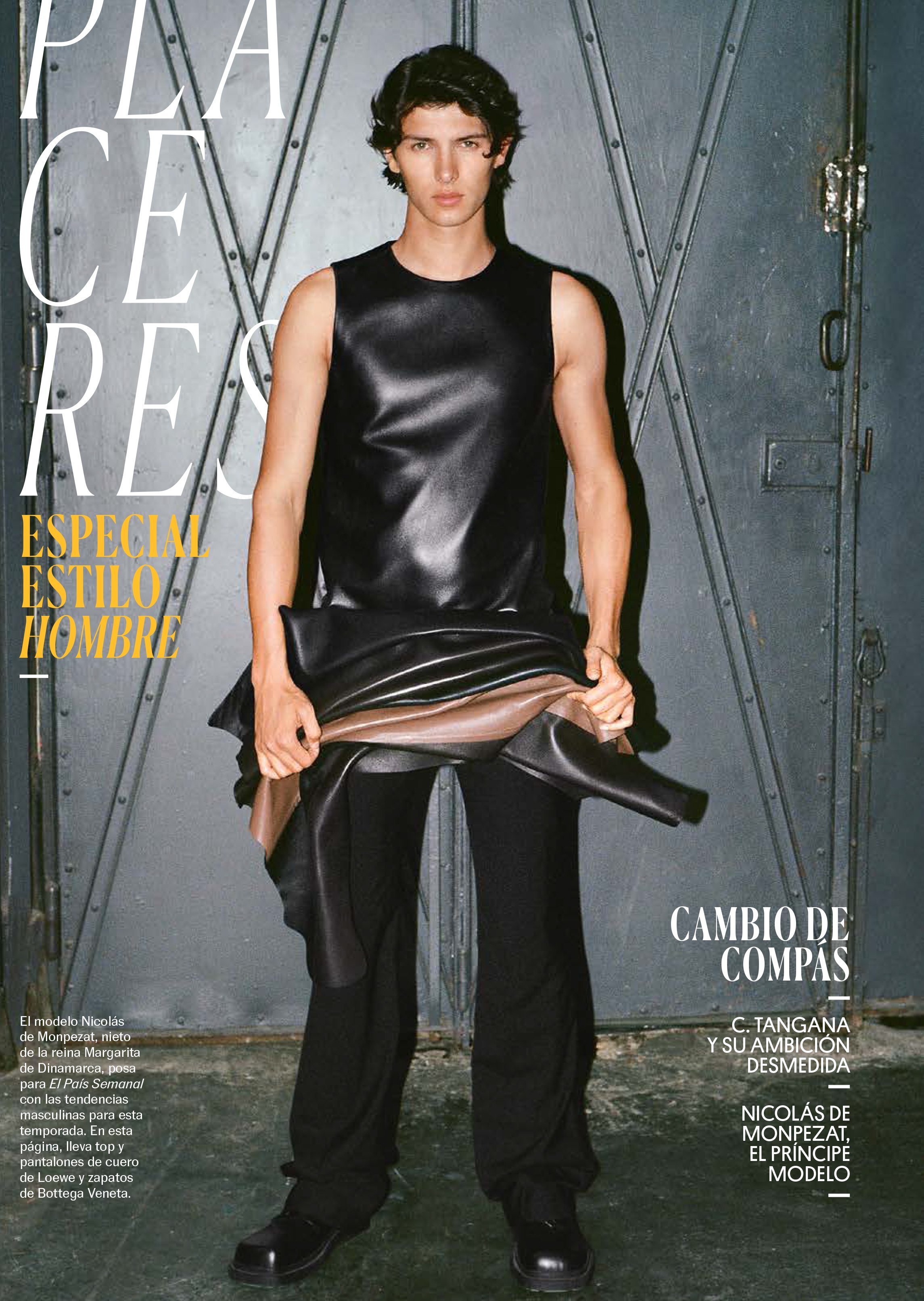 Male fashion model poses in a black sleeveless top, black pants, and black dress shoes against a gray metal background.