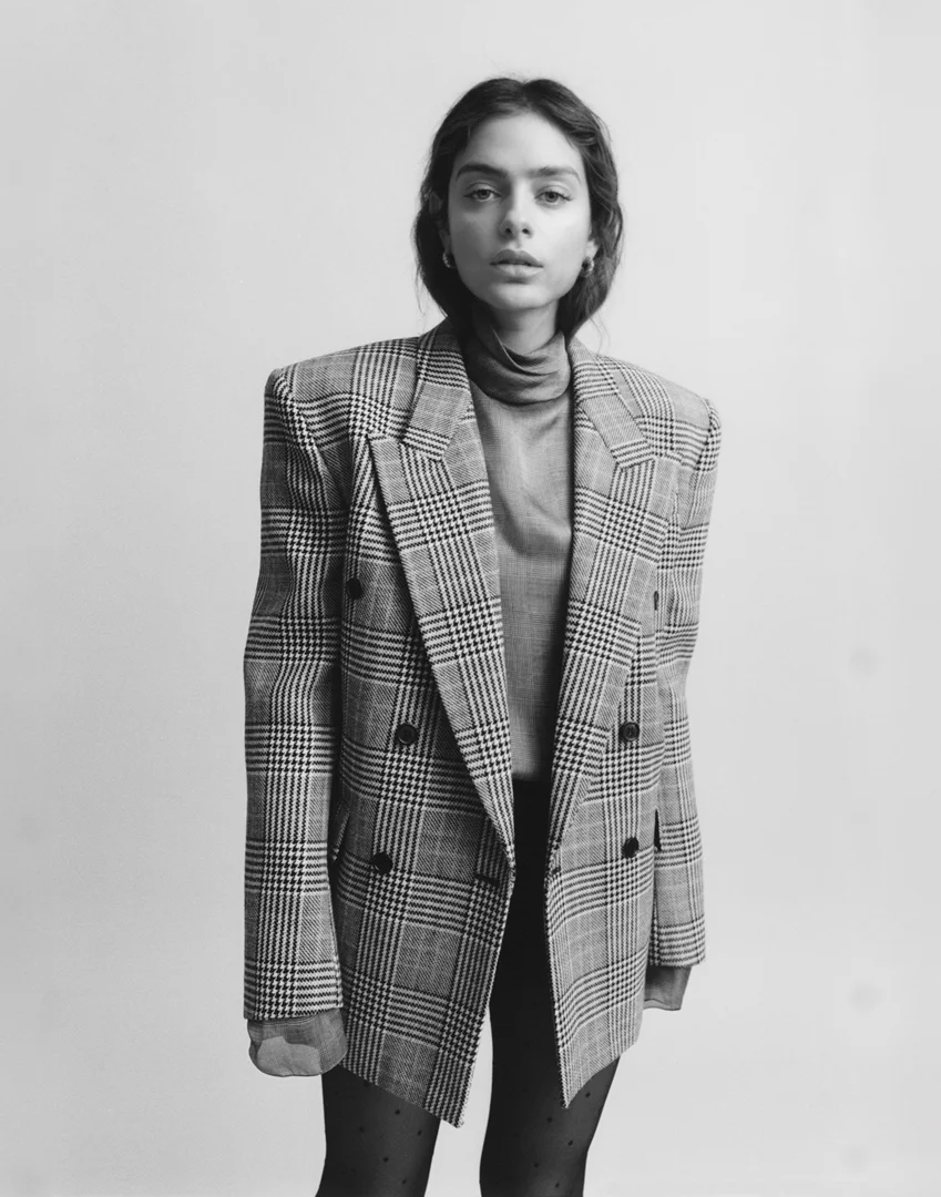 Monochrome portrait of a young adult woman wearing a patterned blazer and turtleneck in a photo studio setting, looking at the camera.