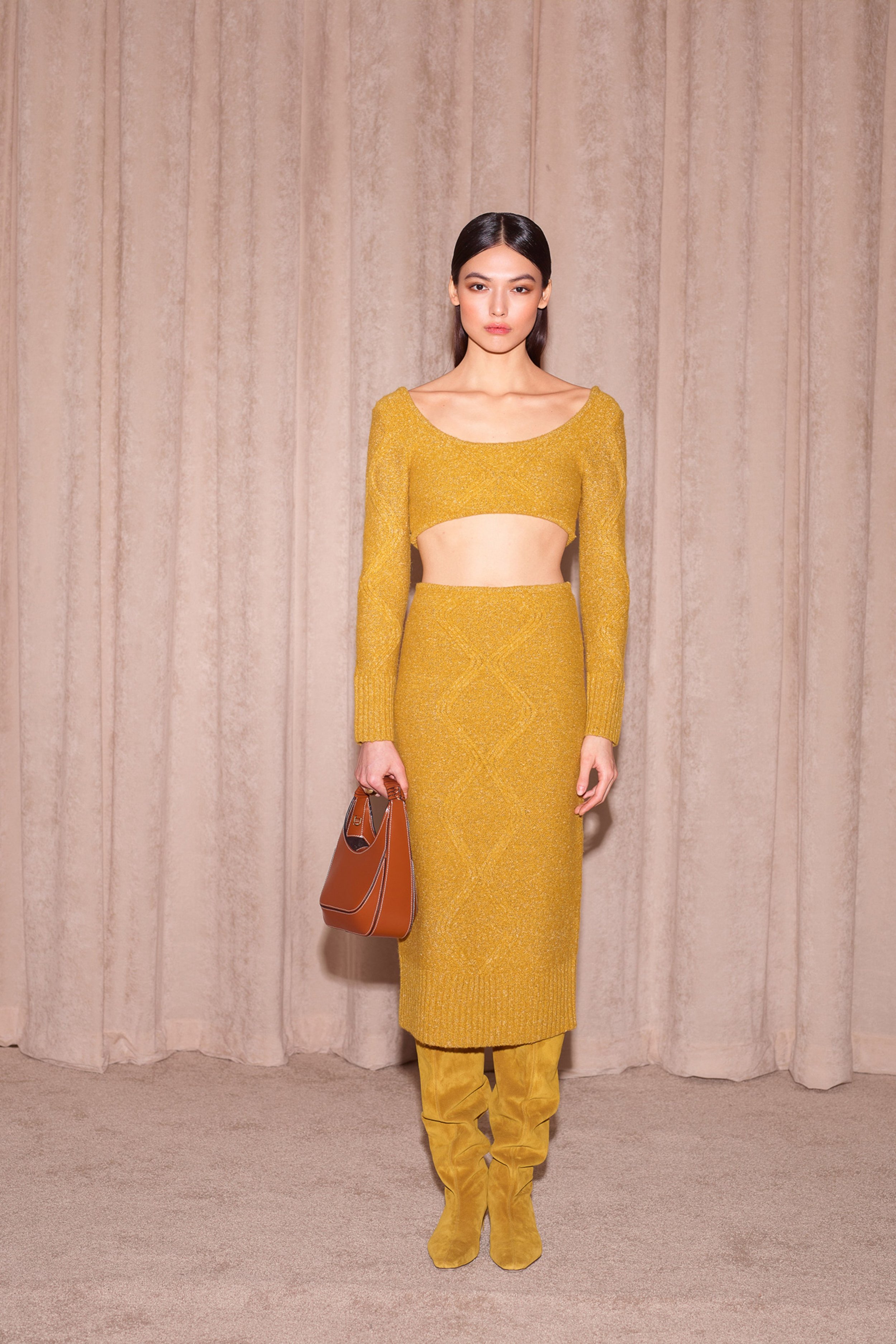 Full-body shot of a woman in a yellow knit skirt and top, accessorized with a brown handbag, standing in front of a neutral-toned curtain.
