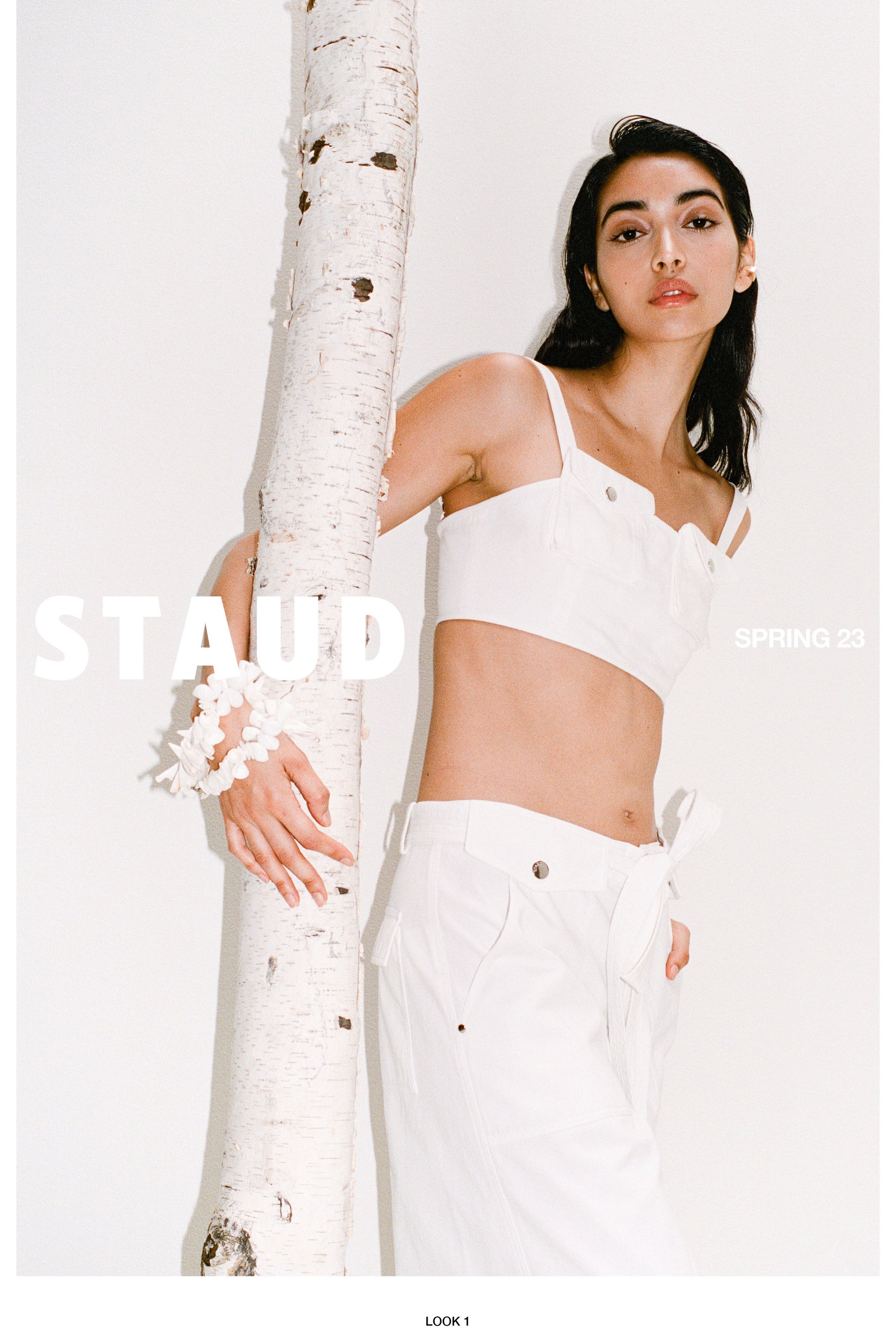Fashion shot of a woman posing in white top and pants next to a birch tree against a white backdrop in natural lighting.