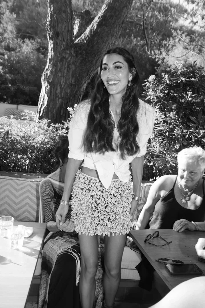 Monochrome image of a smiling woman standing outdoors wearing a tied white blouse and an embellished miniskirt.