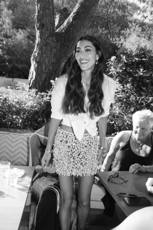 Monochrome image of a smiling woman standing outdoors wearing a tied white blouse and an embellished miniskirt.