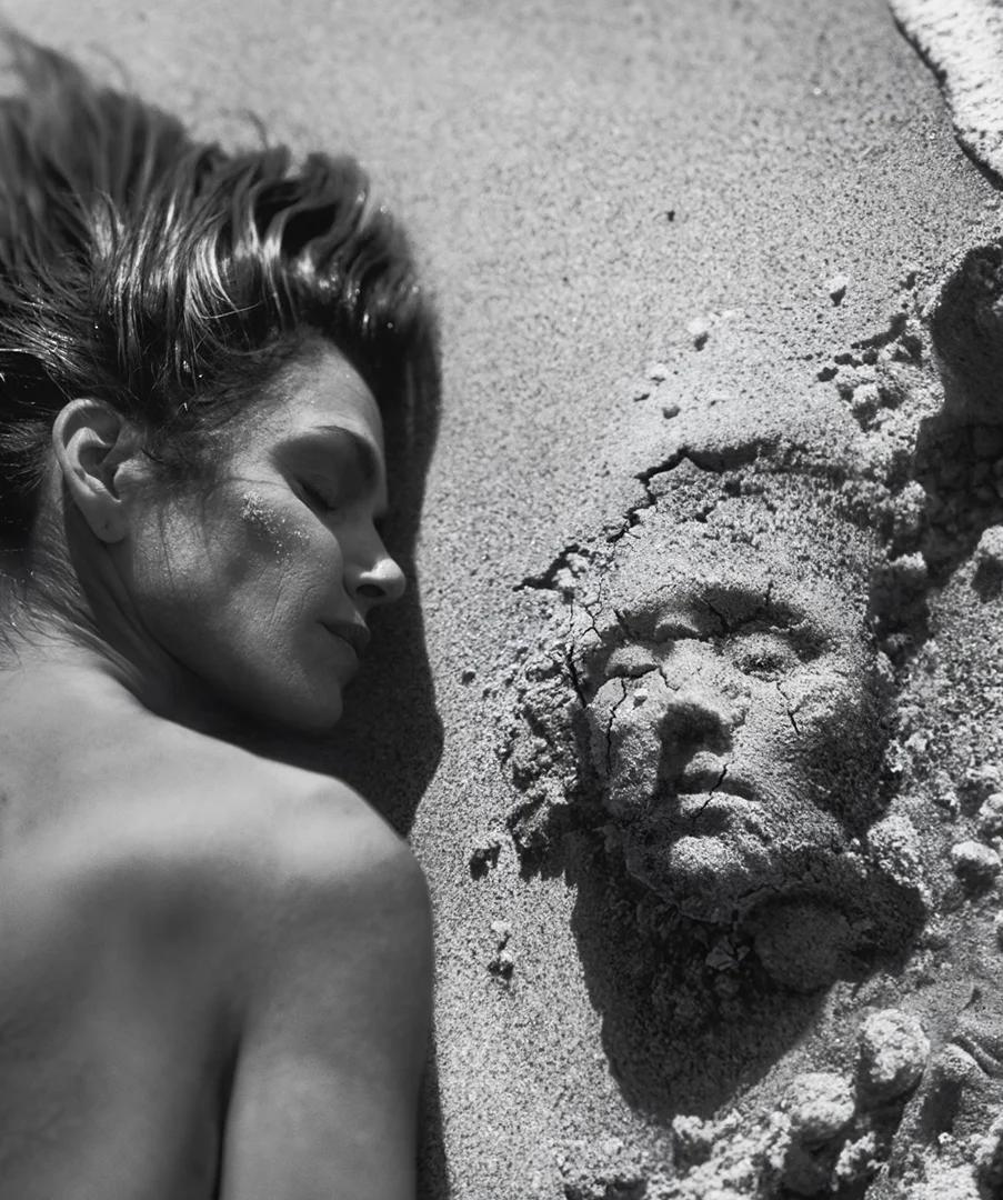 Black and white portrait of a woman lying on a sandy beach next to a human face sculpted in the sand, eyes closed.