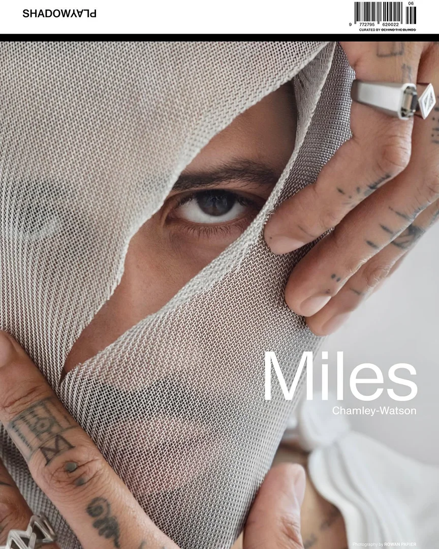 Close-up of a model's face partially covered with mesh fabric, featuring eye contact, tattooed hands, silver rings, and magazine text.