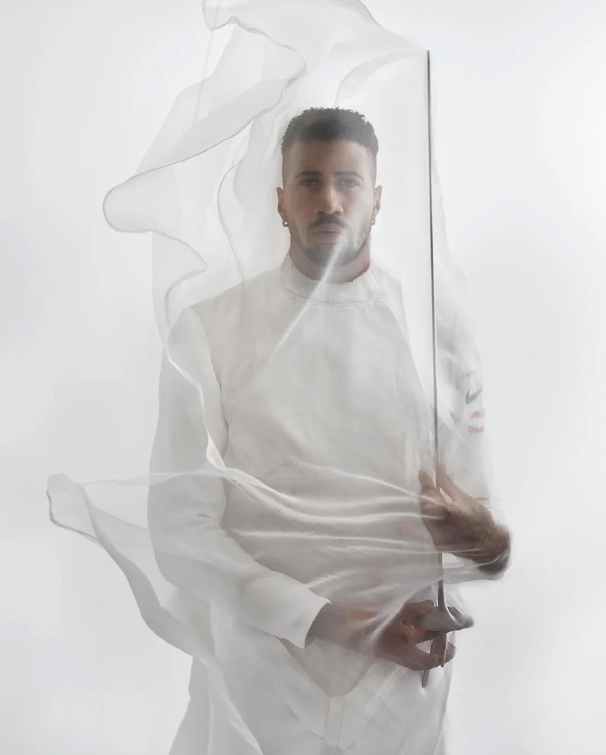 Portrait of adult male fencer in white uniform holding sword, partially obscured by sheer white fabric, in a bright photo studio.