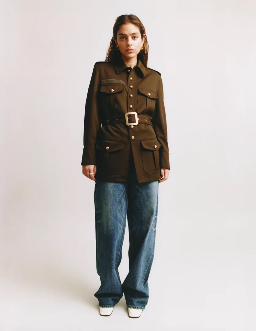 Model wearing an olive green military-style jacket with a belted waist and blue jeans against a light background.