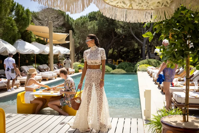 Woman in a lace two-piece outfit stands near a pool, with other people relaxing in the background, creating a resort-style setting.