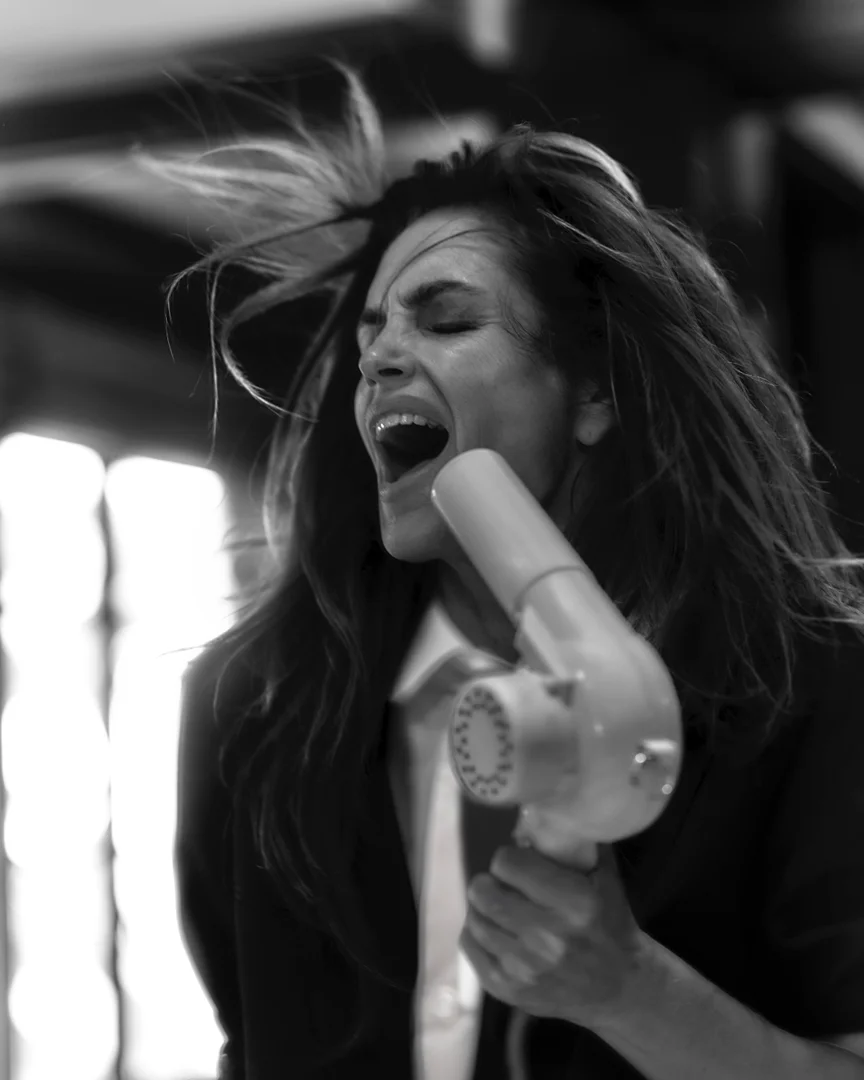 A monochrome portrait captures a woman yelling into a vintage hairdryer, her hair in motion, set against an out-of-focus neutral backdrop.