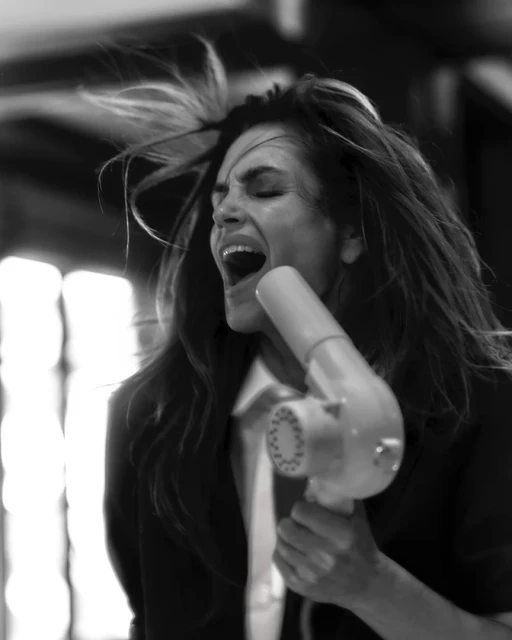 A monochrome portrait captures a woman yelling into a vintage hairdryer, her hair in motion, set against an out-of-focus neutral backdrop.
