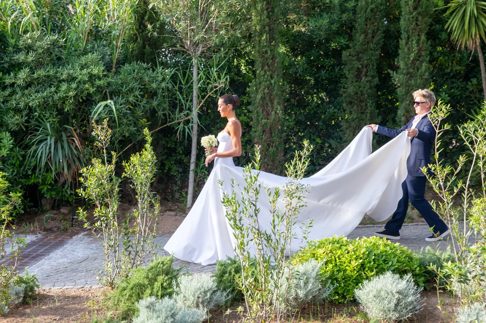 Bride in a white gown walks on a cobblestone path in a garden while a person in a suit holds the long train of her dress.