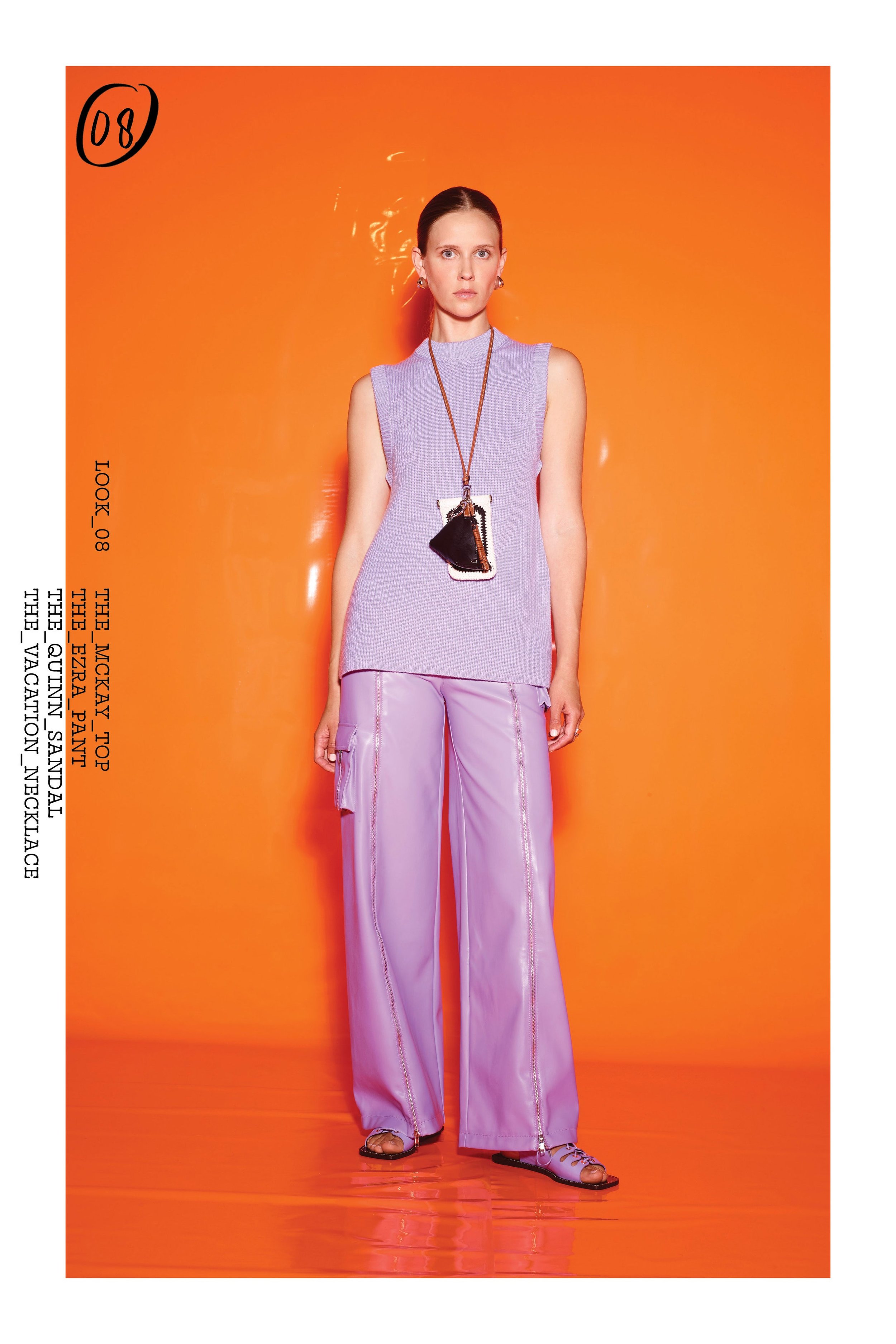 A young adult female fashion model poses in a sleeveless lilac top and wide leg pants against an orange background in a studio.