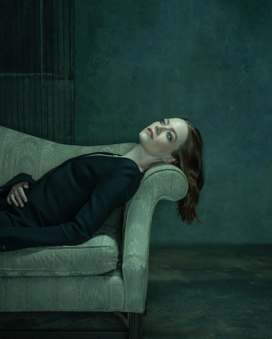 Woman with red hair lounges elegantly on a vintage green sofa against a dark, textured wall in a moody portrait setting.