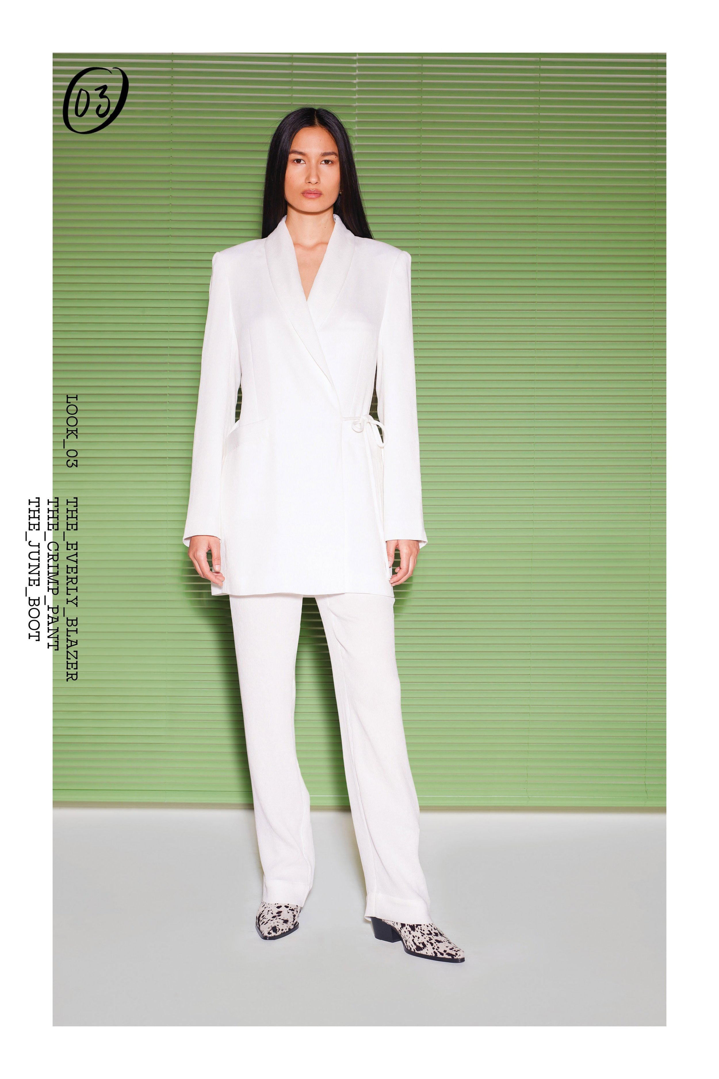 Fashion model poses in a white blazer and pants against a green backdrop in a studio setting, showcasing modern elegance.