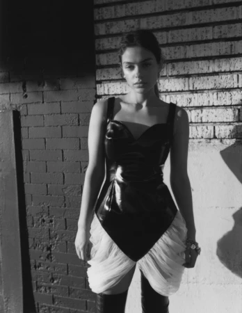 Model wearing a black glossy bodysuit and white ruffled skirt standing against a brick wall.