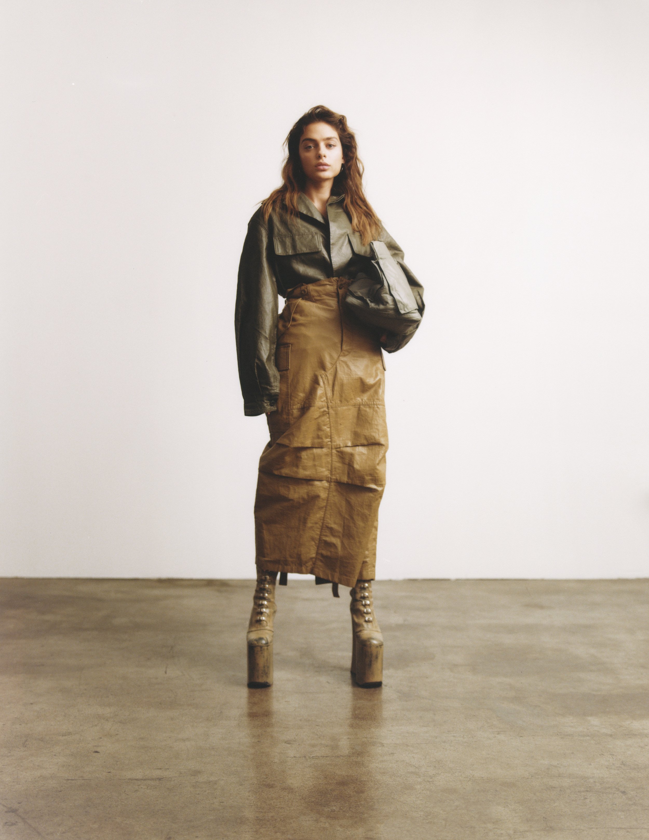 A stylish woman poses in an olive-green jacket, tan skirt, and platform boots against a stark white background in a fashion editorial shot.