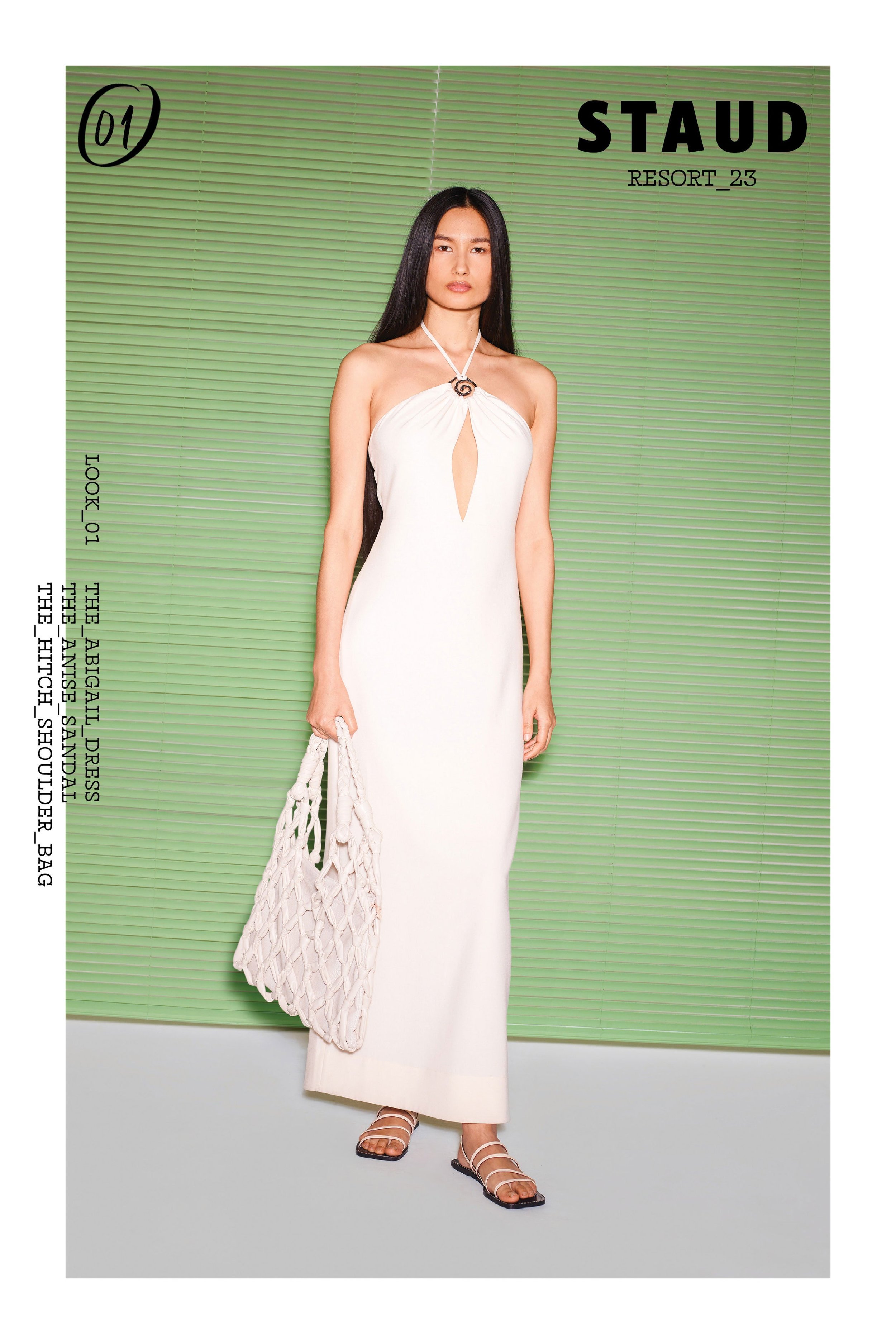 Woman poses in a white halter dress with a keyhole cutout, holding a net bag against a green backdrop for a fashion editorial.