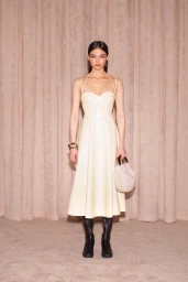 A female model wearing a cream midi dress and black boots holding a small handbag against a patterned fabric background.