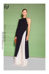 Model standing in a black asymmetrical tunic and white wide-leg trousers holding a brown clutch against a green slatted backdrop.