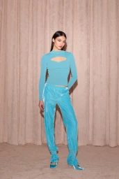 A female model wearing a turquoise cutout top and matching blue velvet trousers standing against a pleated curtain backdrop.
