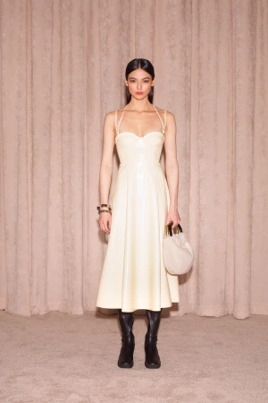 Model in a cream spaghetti strap midi dress holding a small light handbag wearing black boots against a textured curtain backdrop