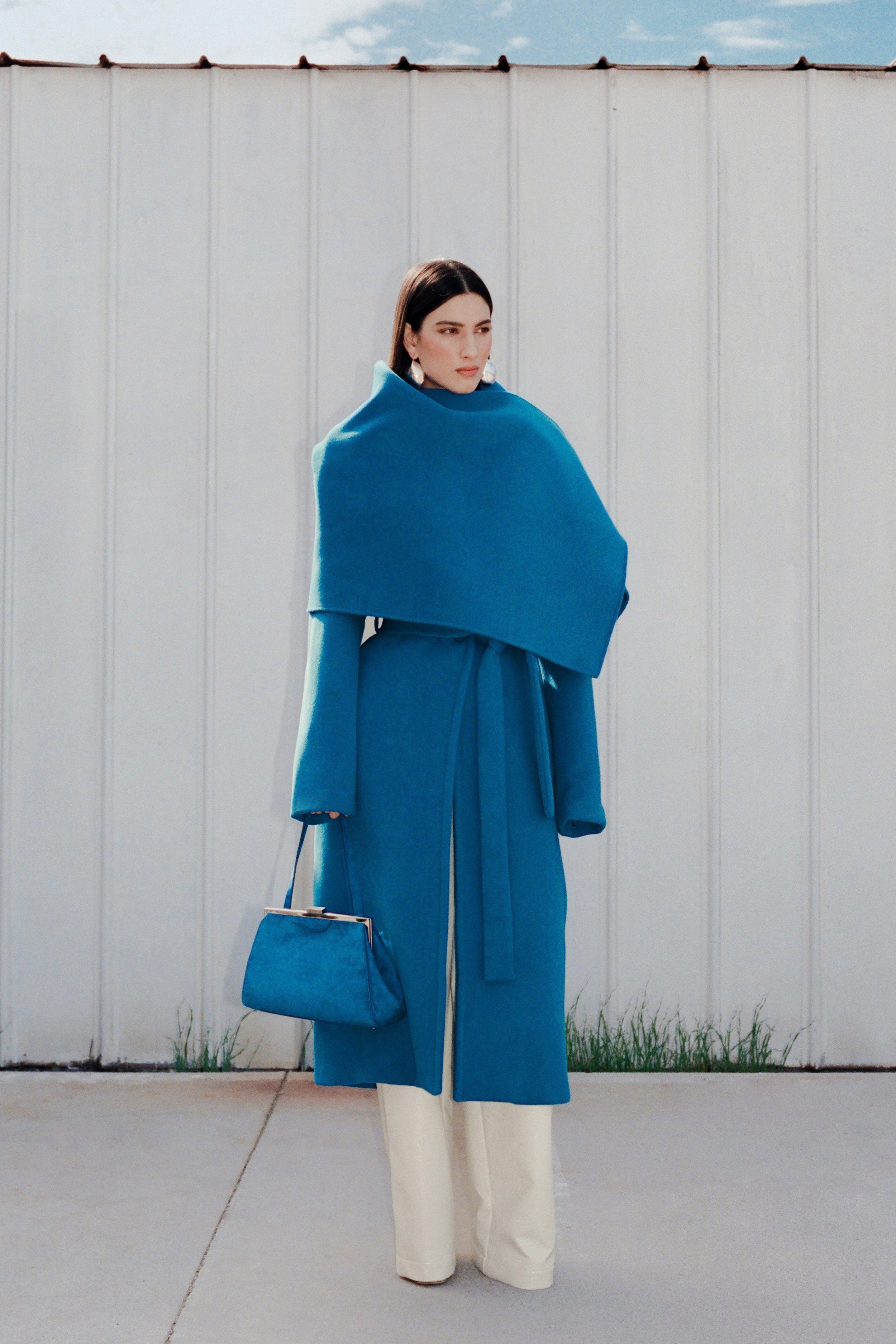 A woman in a long blue coat stands on a concrete surface near a white wall. She carries a blue handbag that complements the coat.