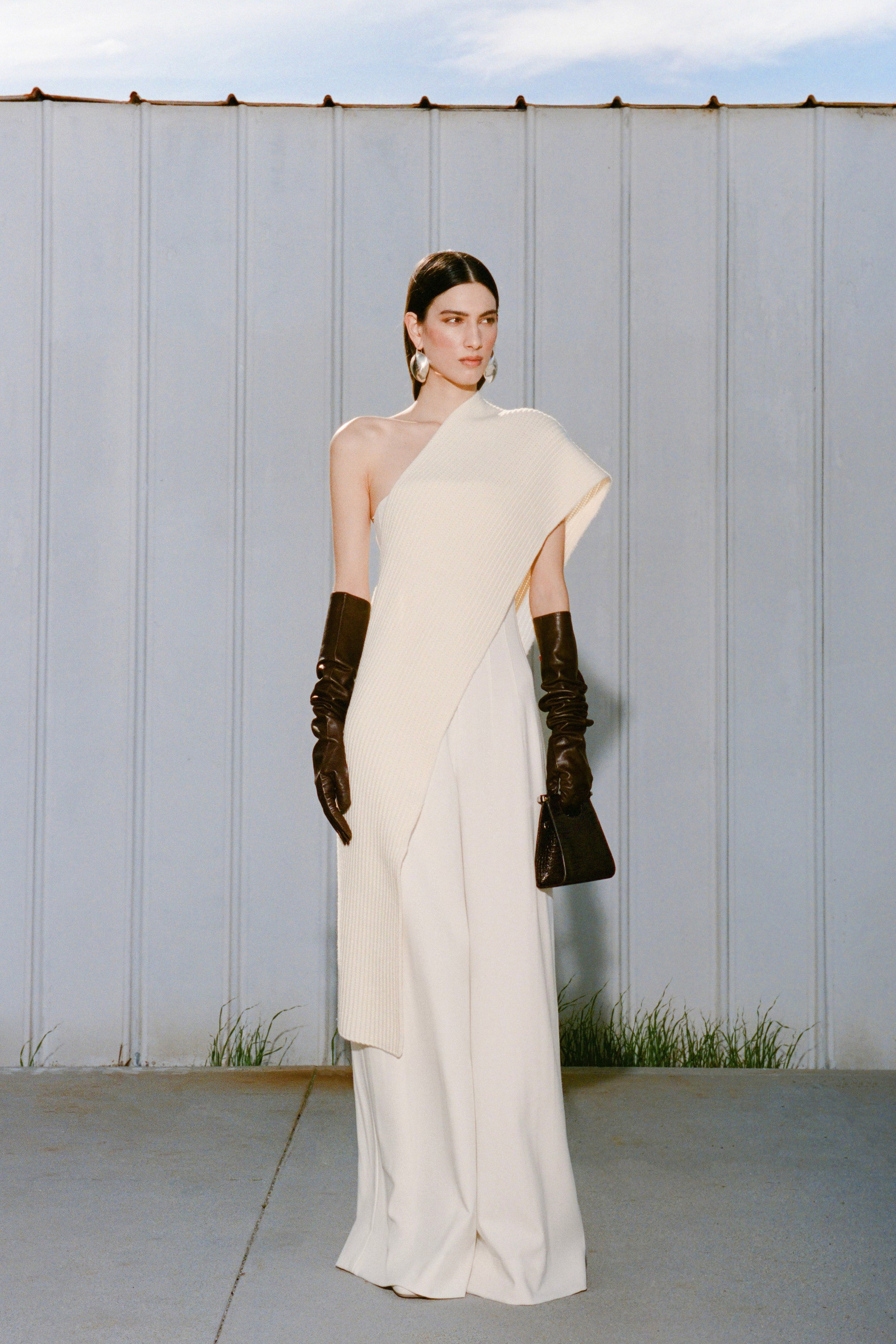 Elegant woman poses in a chic white one-shoulder gown with long gloves and a structured black handbag against a neutral backdrop.