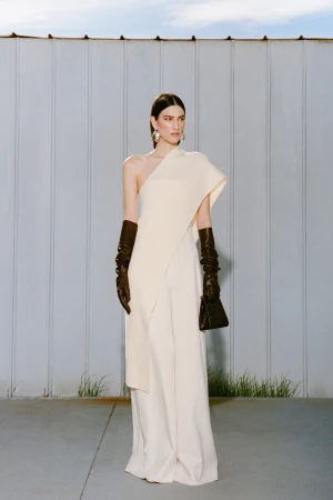 Model wearing a cream asymmetric top, white trousers, and black leather gloves standing before a corrugated metal wall