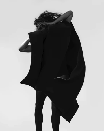 Monochrome fashion portrait of a model wrapped in a structured black coat against a seamless light background.