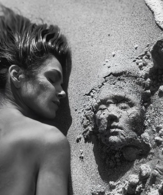 Black and white photograph of a woman lying on a sandy beach next to a sand sculpture of a face with cracked details.