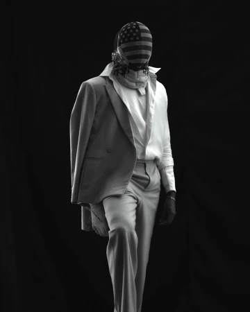 Black and white studio shot of a person wearing a full face covering with an American flag print, suit and gloves, standing.