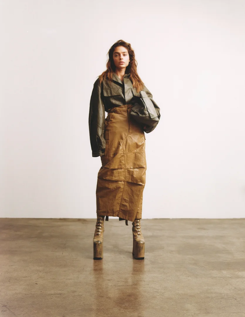 A woman in an olive-green collared shirt, tan skirt, and platform boots stands in a photo studio, holding an olive-green bag.