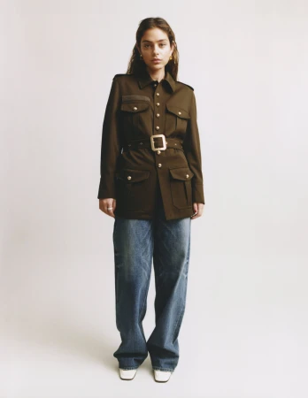 Model wearing a brown utility jacket with a belt and blue jeans against a plain white background