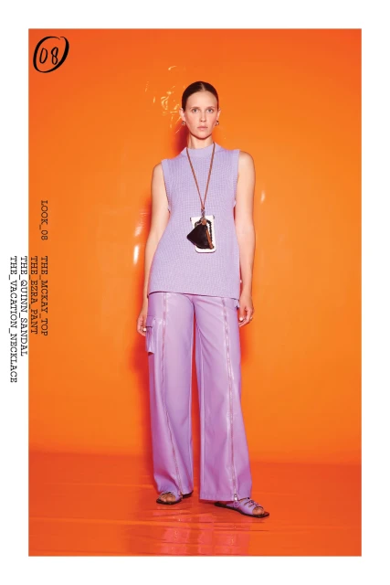 Fashion editorial shot of a woman in a lavender top and pants with a large pendant necklace against a bright orange backdrop.
