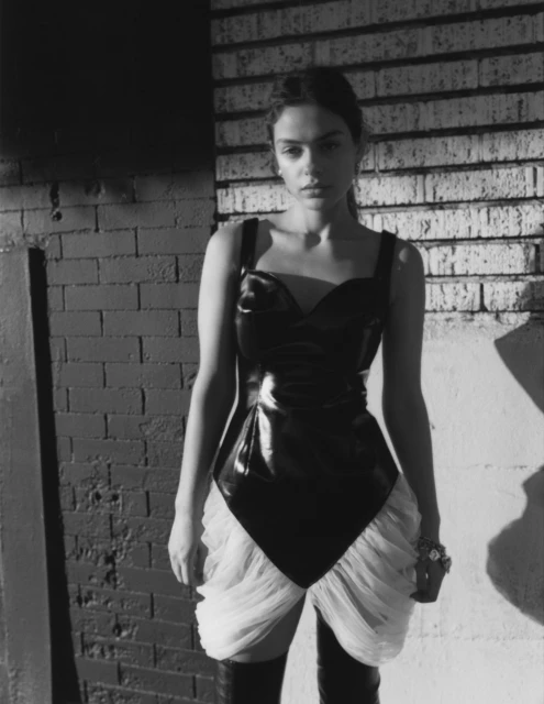 Monochrome portrait of a woman in a sleek black bodysuit and thigh-high boots against a brick wall with hard lighting.