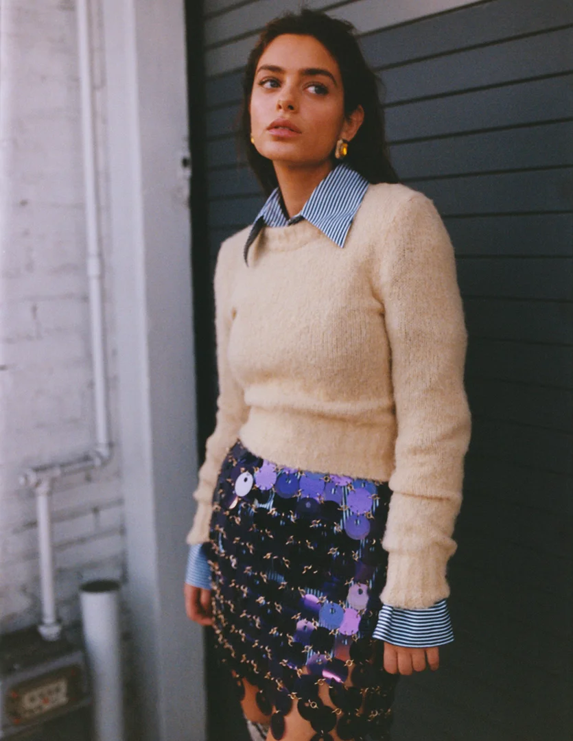 Fashion portrait of a brunette woman wearing a beige sweater and sequined skirt against a dark wall. Natural light, medium shot.
