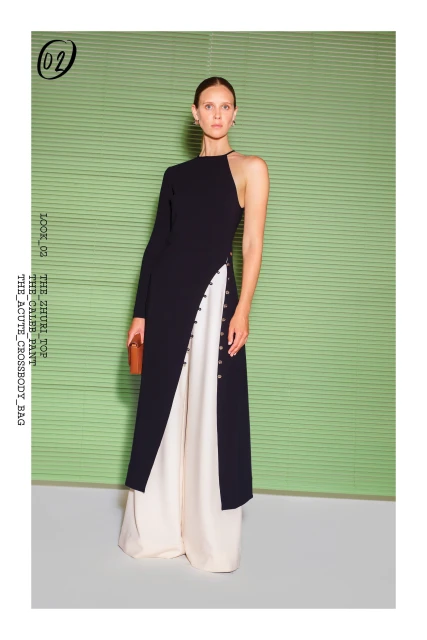 Fashion model poses in studio wearing black one-shoulder dress with side slit and white pants against a green backdrop.