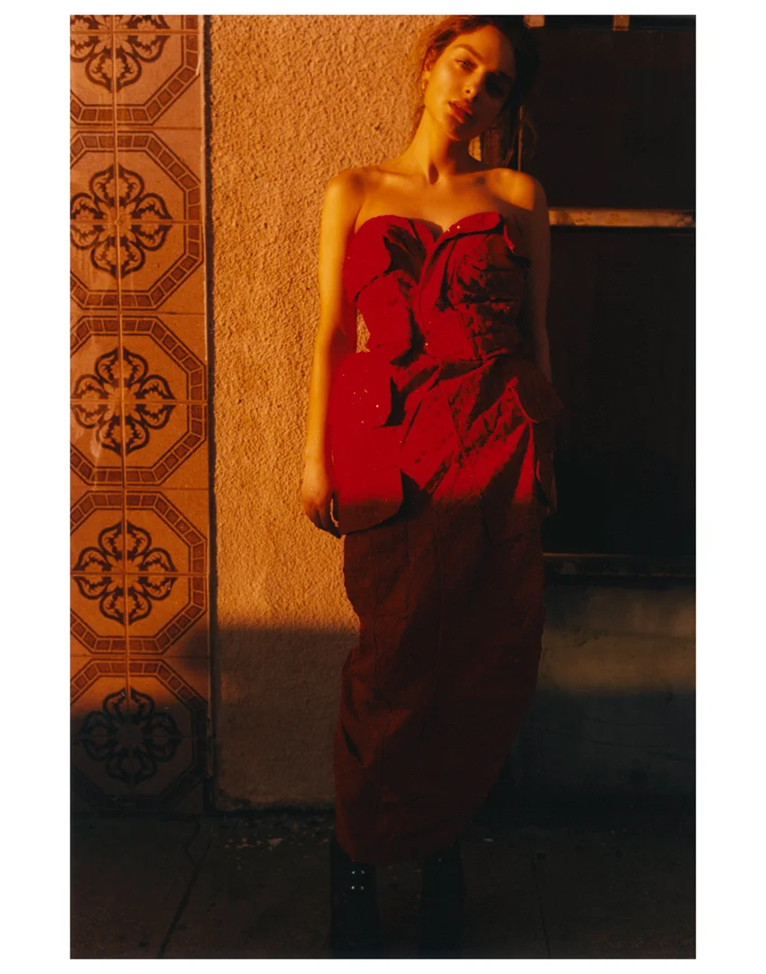 A light-skinned adult female poses in a long, strapless red dress against a textured wall in an urban setting with warm lighting.