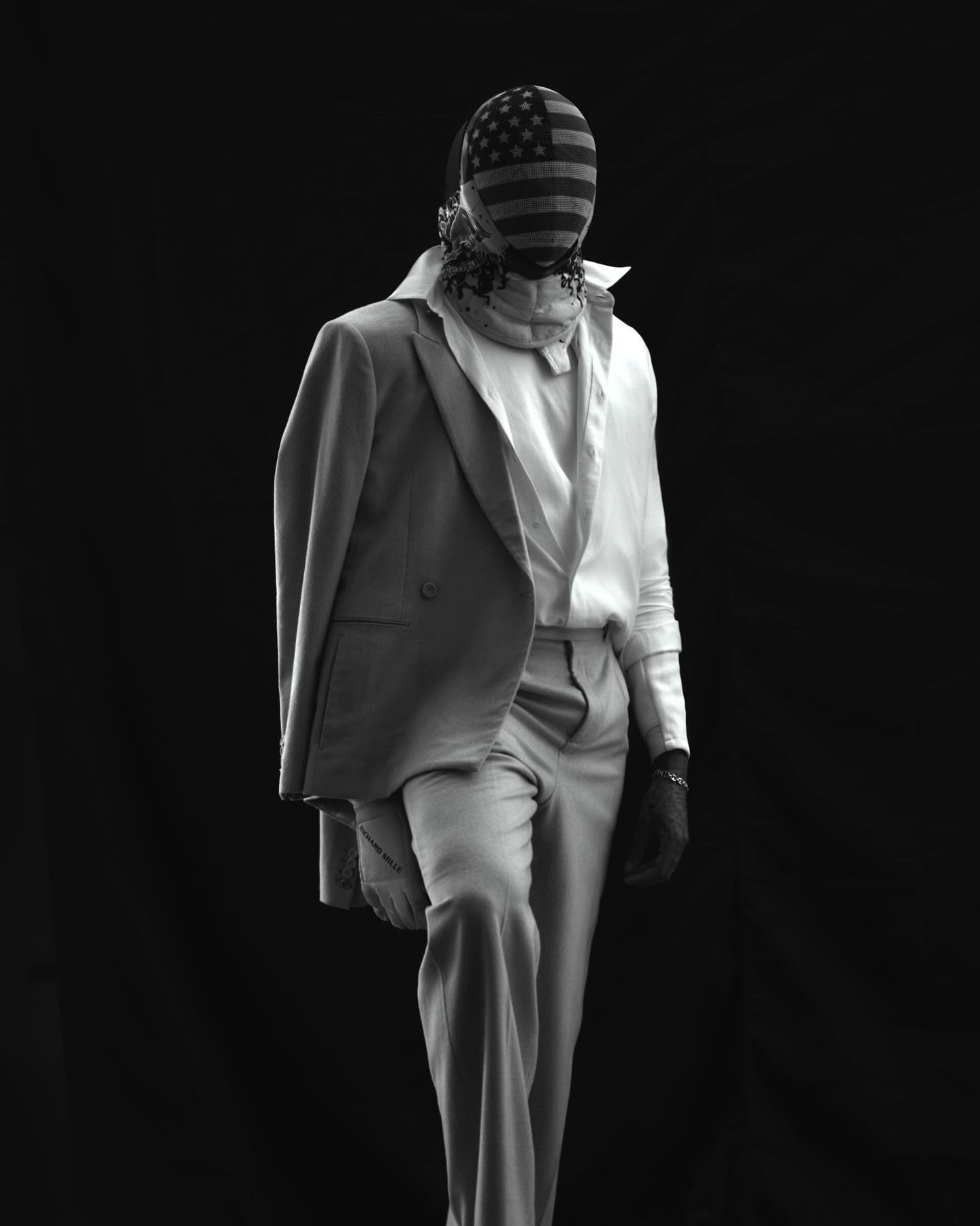 Person in suit with USA flag balaclava stands against a black backdrop, shot in black and white with soft, diffused studio lighting.