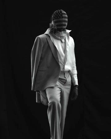 Person in suit with USA flag balaclava stands against a black backdrop, shot in black and white with soft, diffused studio lighting.