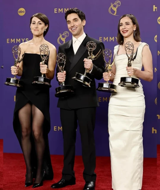 Group portrait of three smiling adult actors holding Emmy awards on a red carpet in front of a purple branded background.