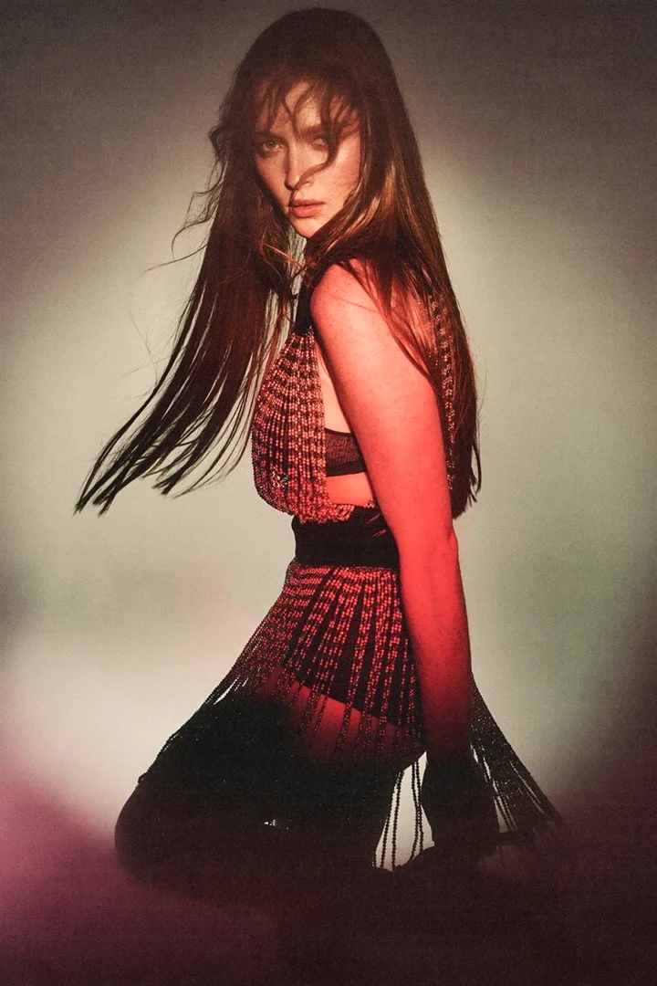 Portrait of a woman with long brown hair wearing a fringed dress in a studio setting with dramatic lighting, captured in a medium shot.