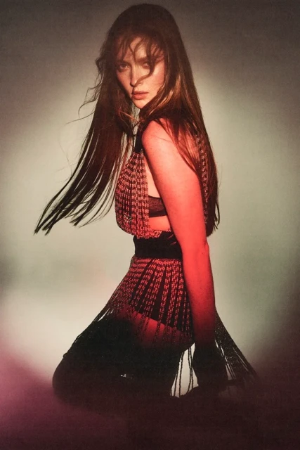 Portrait of a woman with long brown hair wearing a fringed dress in a studio setting with dramatic lighting, captured in a medium shot.