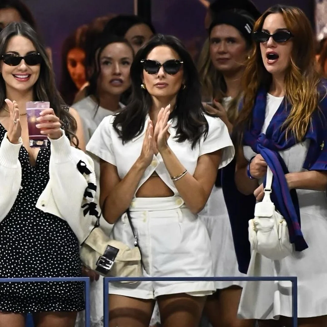 Three women in sunglasses at an outdoor event. One holds a drink, another claps, and another wears a blue scarf. Fashionable attire.