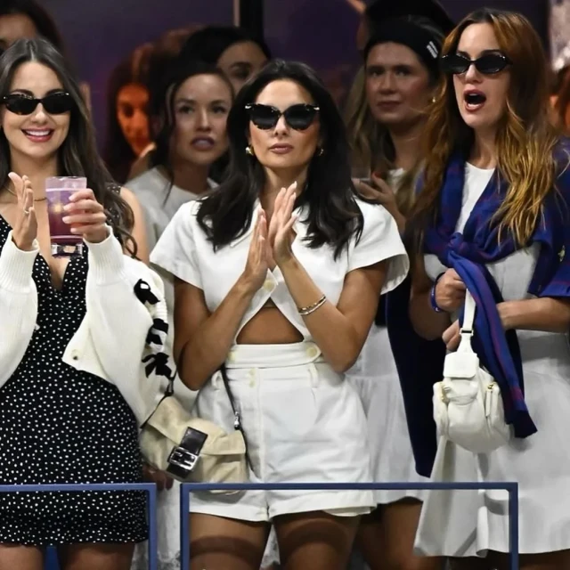 Three women in sunglasses at an outdoor event. One holds a drink, another claps, and another wears a blue scarf. Fashionable attire.