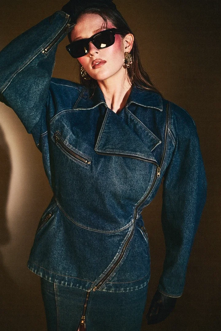 Woman poses in studio lighting wearing a blue denim jacket with a visible zipper, complemented by sunglasses and statement earrings.