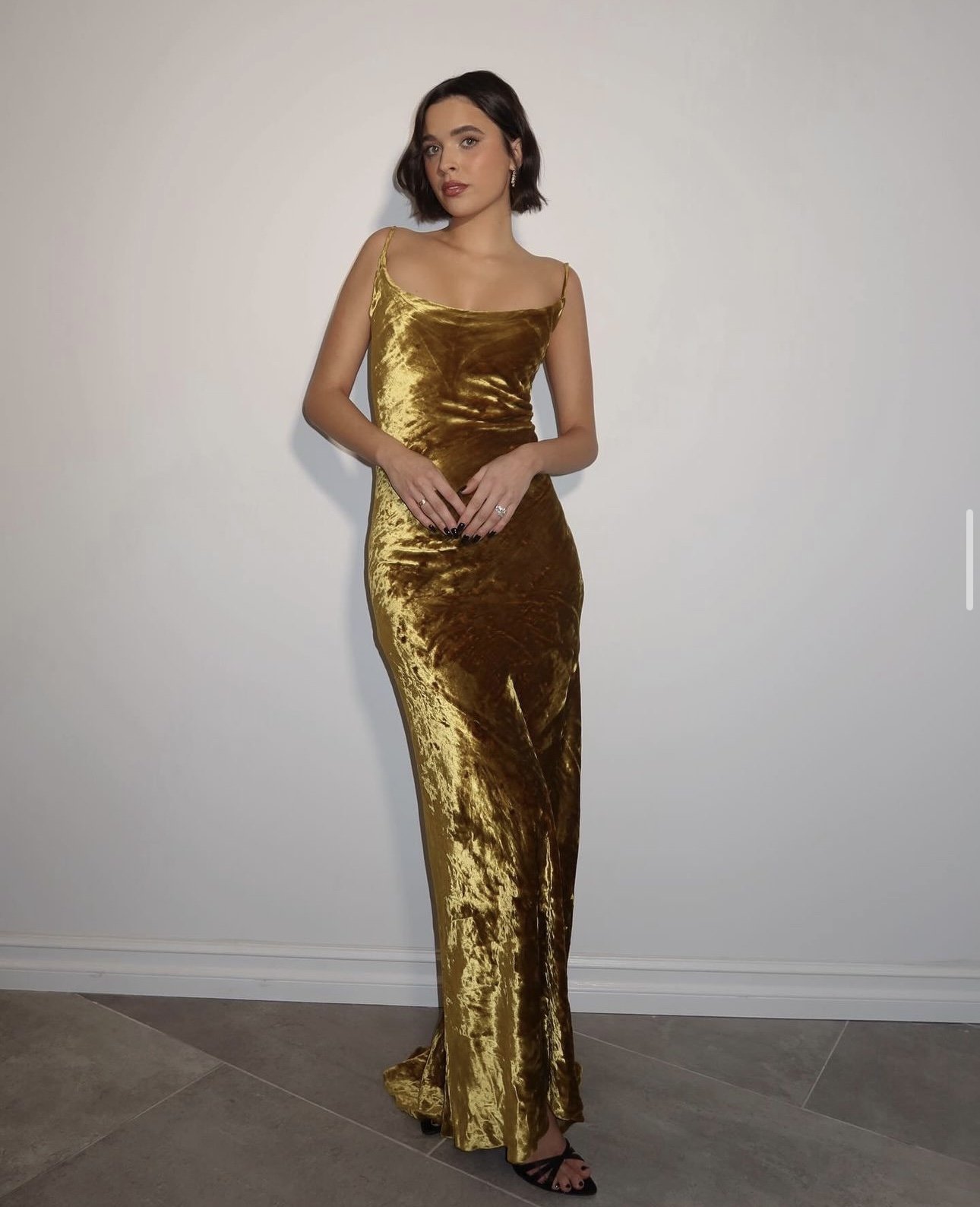 Portrait of a woman in a gold velvet dress. She is indoors with neutral lighting, posing against a white wall and grey stone floor.
